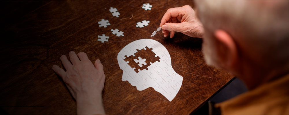 Primer plano de una persona mayor intentando completar un rompecabezas con forma de perfil humano; faltan piezas en la zona del cerebro, simbolizando la pérdida de memoria o el Alzheimer.