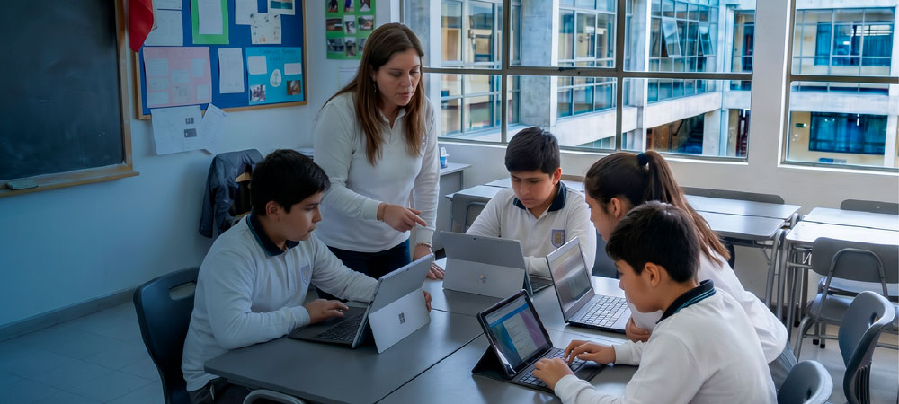 Una profesora de pie asiste a cuatro estudiantes de uniforme, dos niños y dos niñas, que trabajan concentrados con computadores portátiles y tablets en una mesa de clases. Al fondo se ve un gran ventanal que da hacia el patio del colegio.
