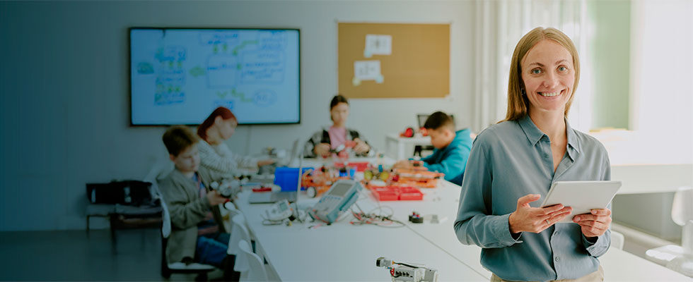 Una profesora sonriente sostiene una tableta en primer plano dentro de un aula de tecnología, mientras un grupo de estudiantes trabaja con proyectos de robótica en el fondo.