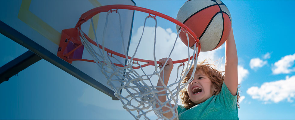 Niño encesta un balón de básquetbol en un aro al aire libre durante una actividad deportiva, con el cielo azul de fondo