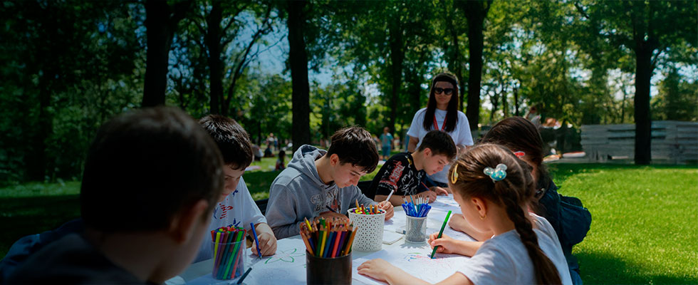 Un grupo de niños y jóvenes sentados alrededor de una mesa al aire libre en un parque rodeado de árboles, participando en un taller de dibujo y pintura con lápices de colores y marcadores bajo la supervisión de un adulto.