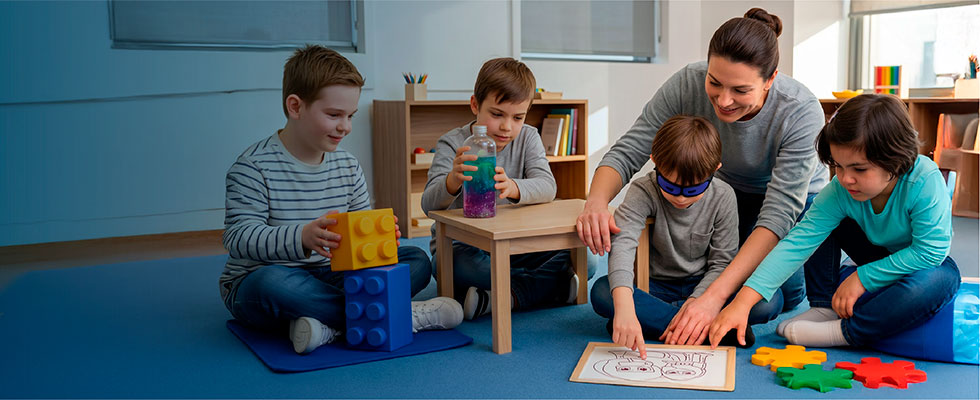 Una educadora junto a un grupo de niños sentados en el suelo de un aula, realizando diversas actividades educativas y sensoriales como armar bloques, explorar frascos sensoriales y utilizar un tablero interactivo.