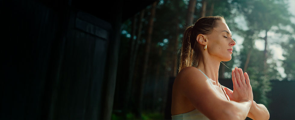 Una mujer en posición de meditación o yoga, con los ojos cerrados y las manos juntas en gesto de oración, en un entorno natural rodeado de árboles.